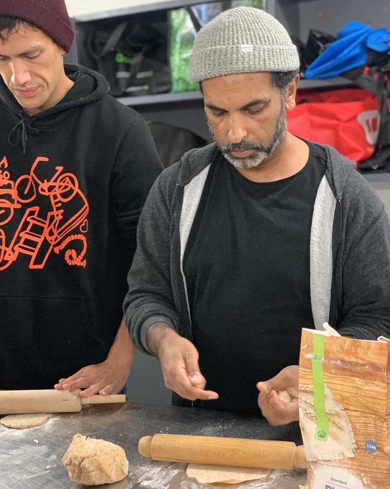 Two men at a kitchen counter rolling and shaping dough with rolling pins and a bag of flour nearby.
