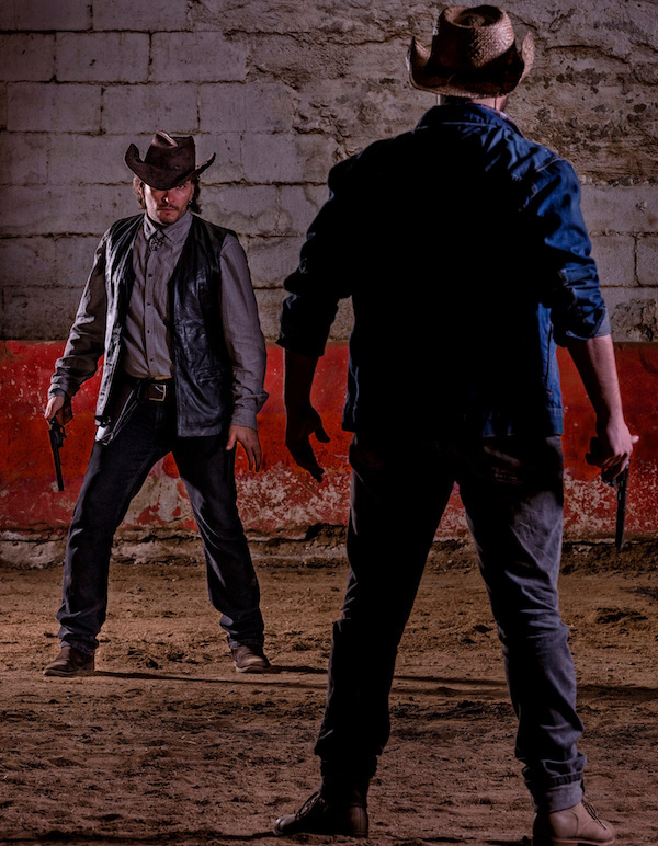 Two cowboys face off in a dusty arena, both holding revolvers; one seen from behind, the other facing forward.