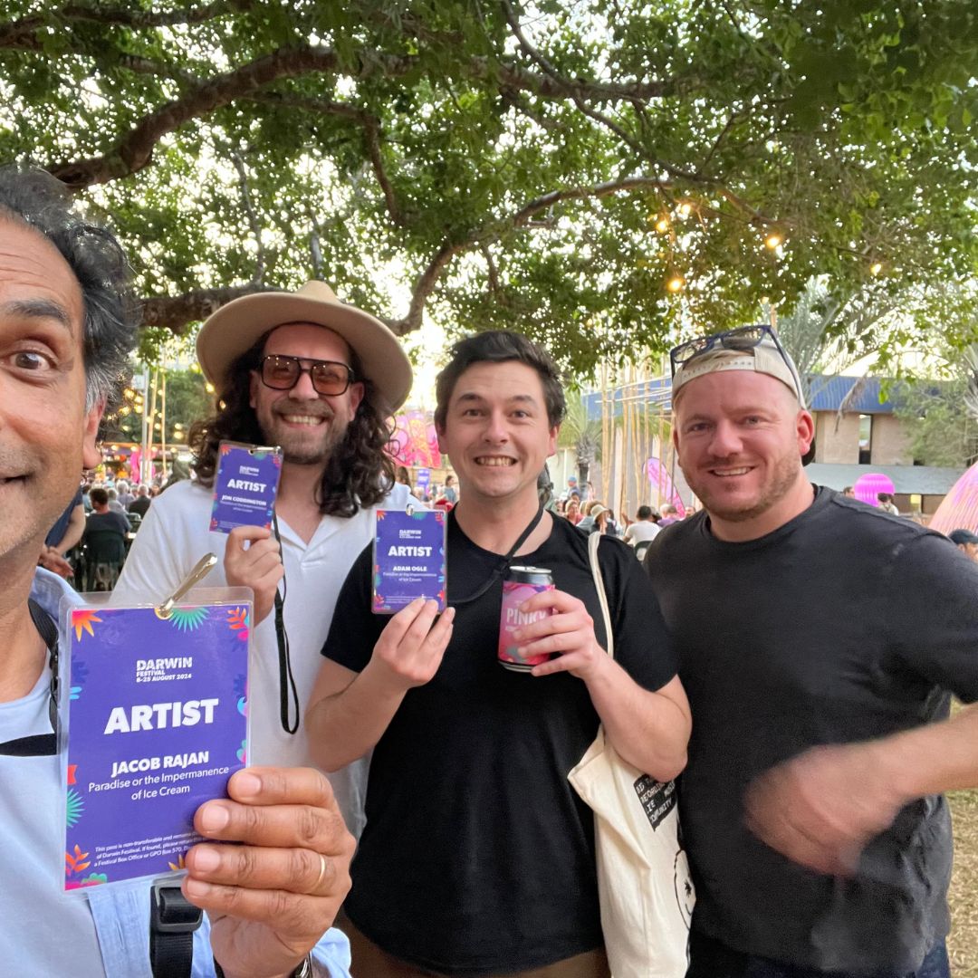 Four smiling men at an outdoor festival under trees, three holding "ARTIST" passes and one holding a canned drink.