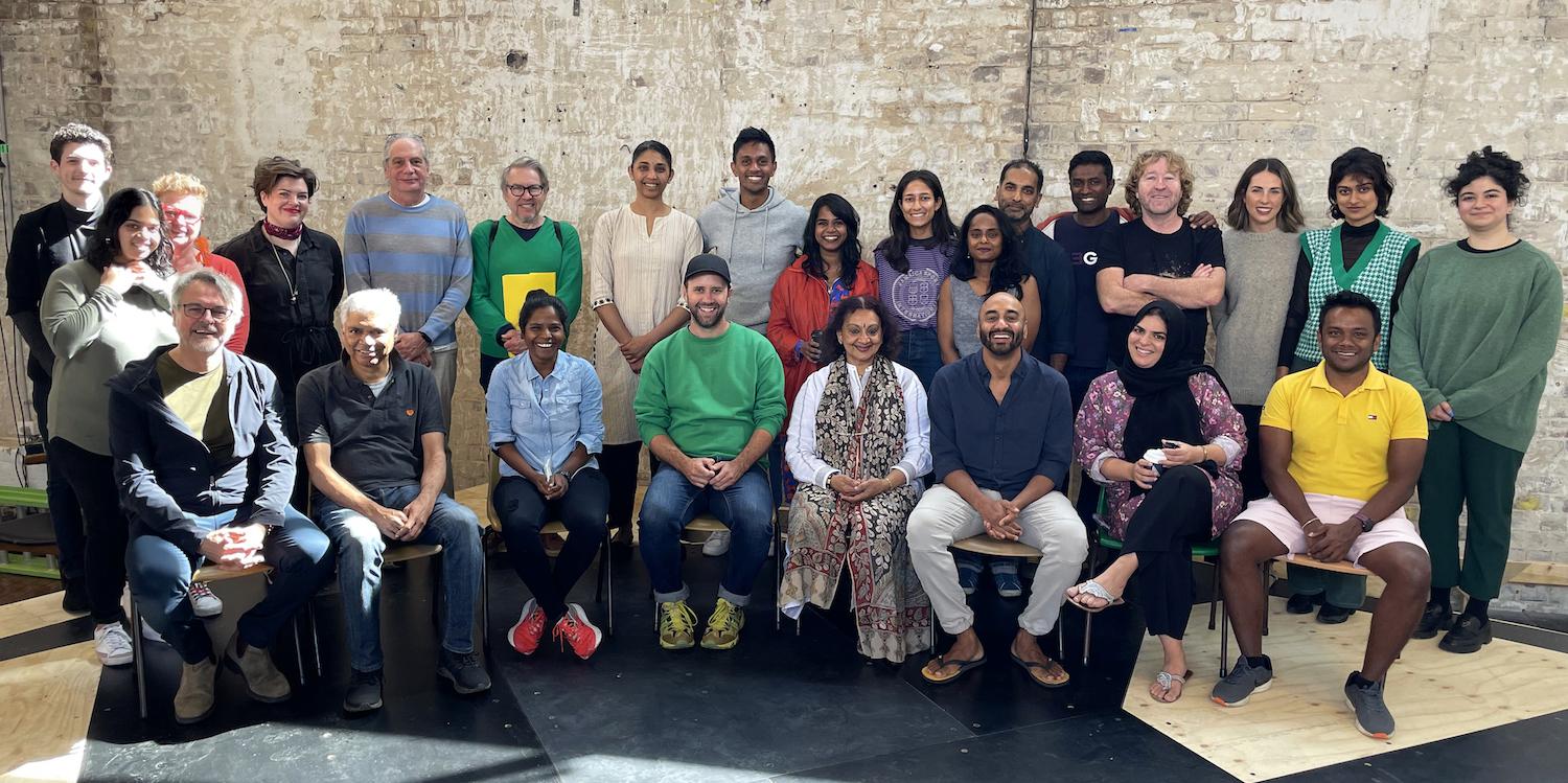 Diverse group of about 25 people, seated and standing, smiling in a bright room against a weathered brick wall.