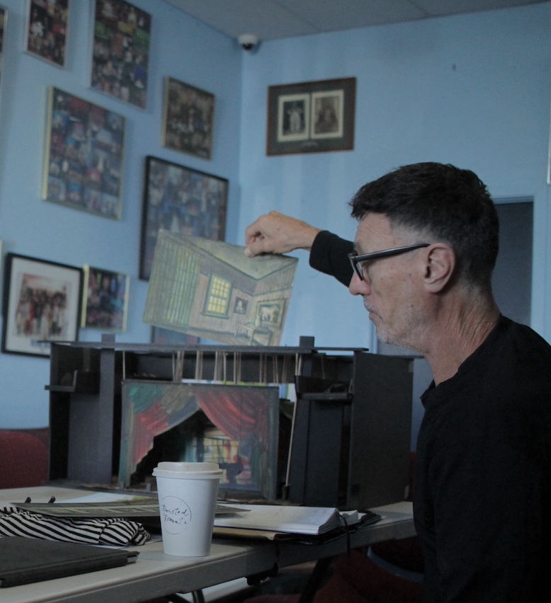 Man with glasses lifts a painted paper house above a miniature stage with red curtains on a table; coffee cup in foreground.