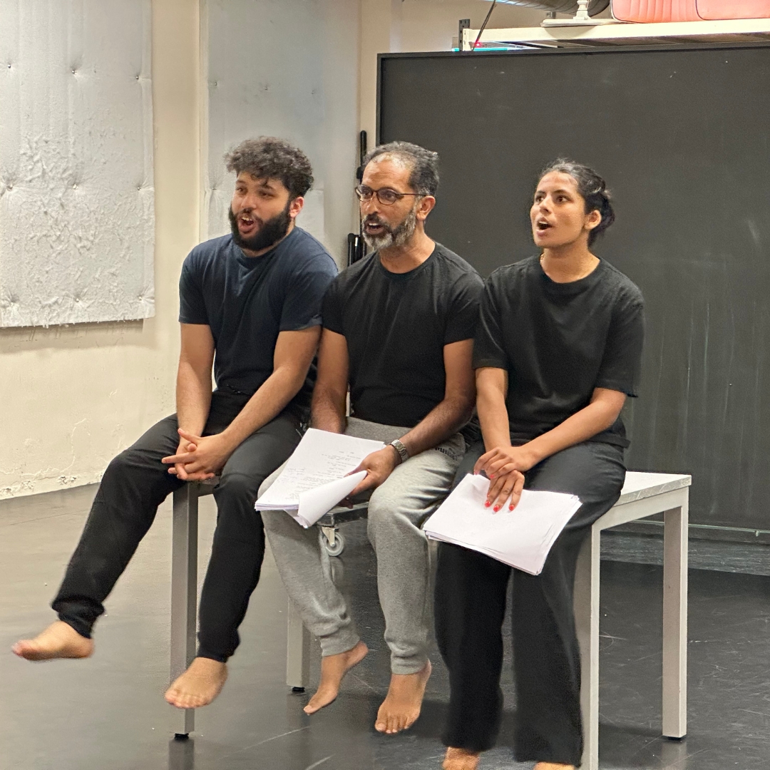Three barefoot performers in black shirts sit on a bench holding scripts, singing or rehearsing in a studio.