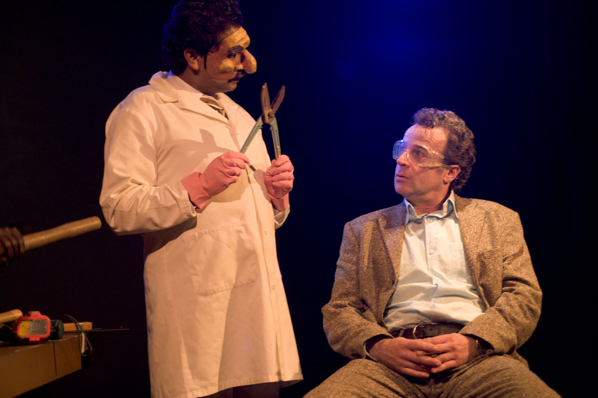 Man in lab coat and rubber gloves with a prosthetic nose holds oversized shears while a seated man in a suit and safety goggles looks up