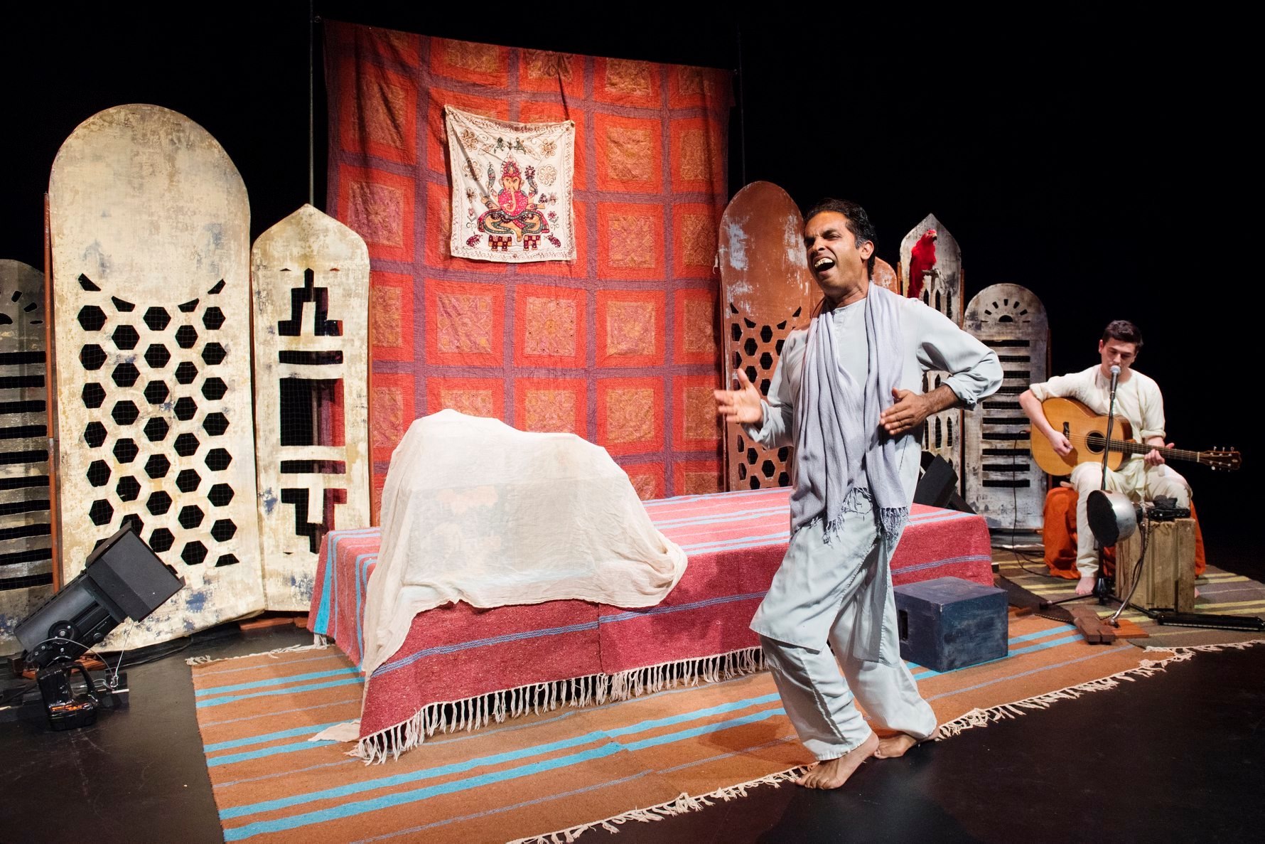 Barefoot male performer in light robes singing energetically on a colorful stage while a seated guitarist accompanies him