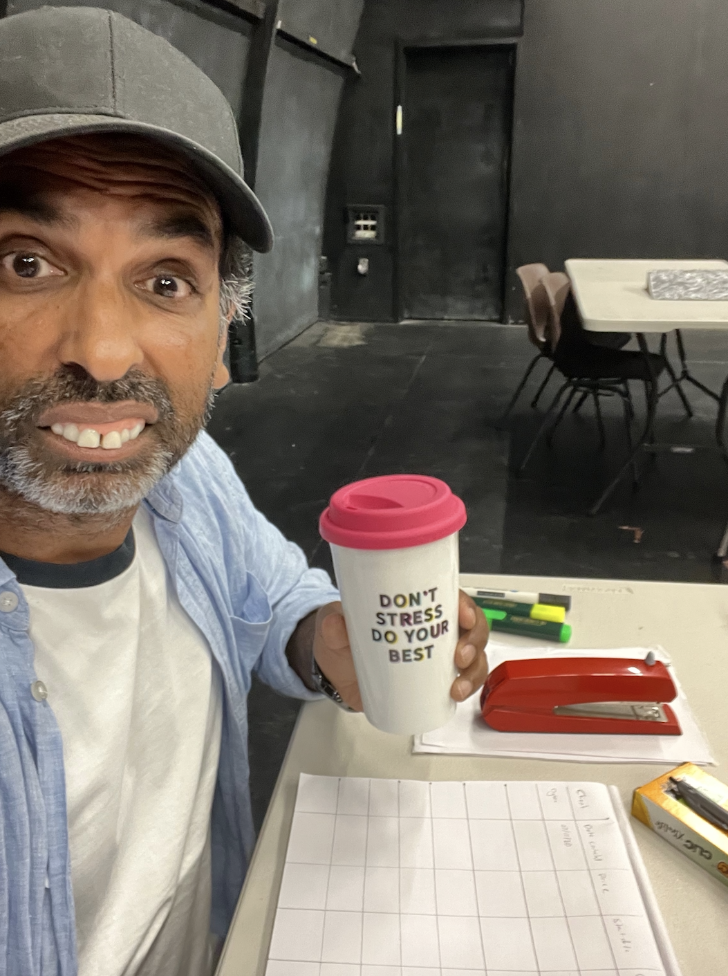 Smiling man wearing a cap holds a white travel mug with a pink lid that reads "DON'T STRESS DO YOUR BEST" at a table.