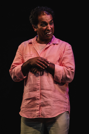 Jake in pink button-down shirt on dark stage, clasping hands and making an animated, expressive face.