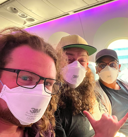Three passengers on an airplane wearing 3M respirator masks; one makes a shaka hand sign under purple cabin lighting.