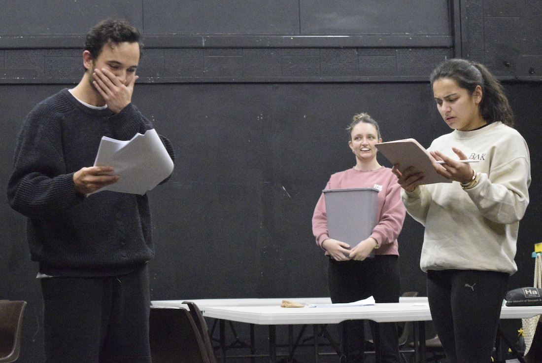 Three actors rehearsing: man covers his mouth while reading a script, woman holding a bin smiles, another reads from a tablet.