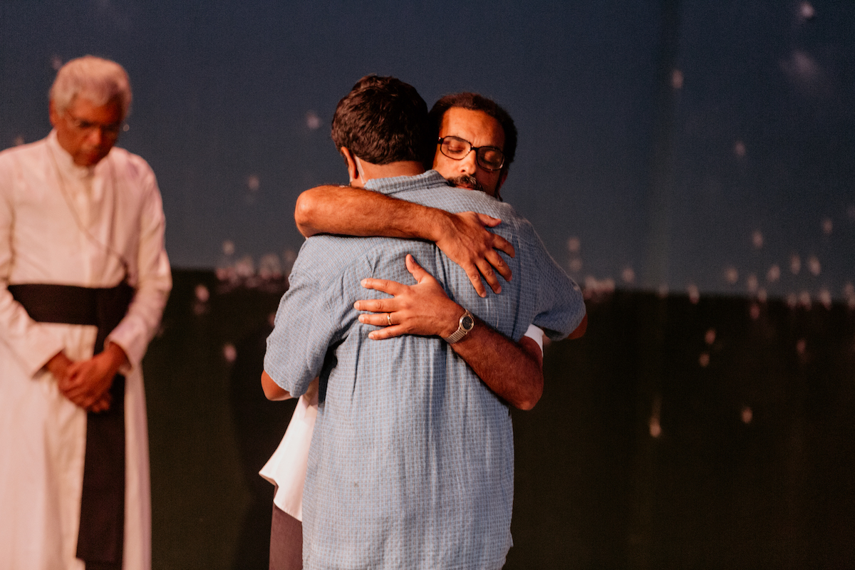 Two men embrace on stage; one wearing glasses and a wristwatch, a robed man stands bowed nearby.