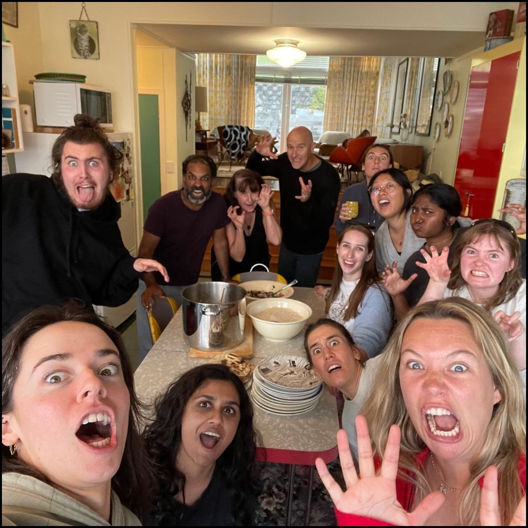 Dozen people in a kitchen selfie making playful scary faces around a table with pots and bowls.