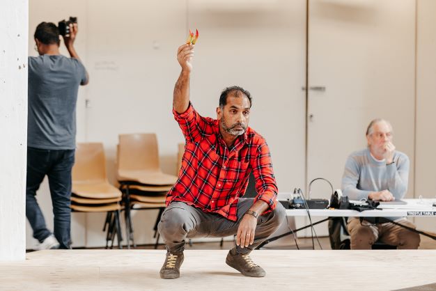 Man in red plaid shirt squatting onstage, holding up small colorful paper shapes while crew members observe and photograph