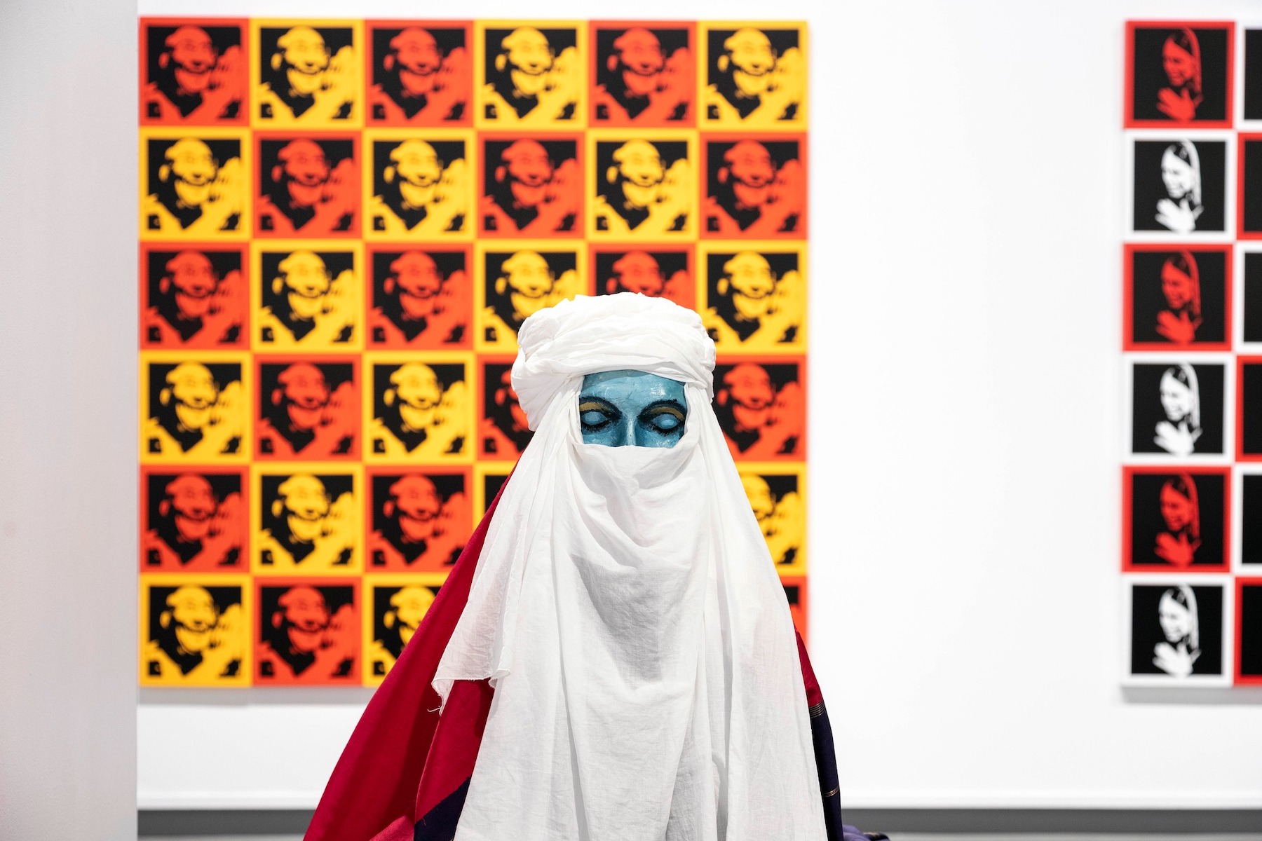 Figure wearing white turban and veil with a blue-painted face and closed eyes, posed before a red-and-yellow pop-art grid.
