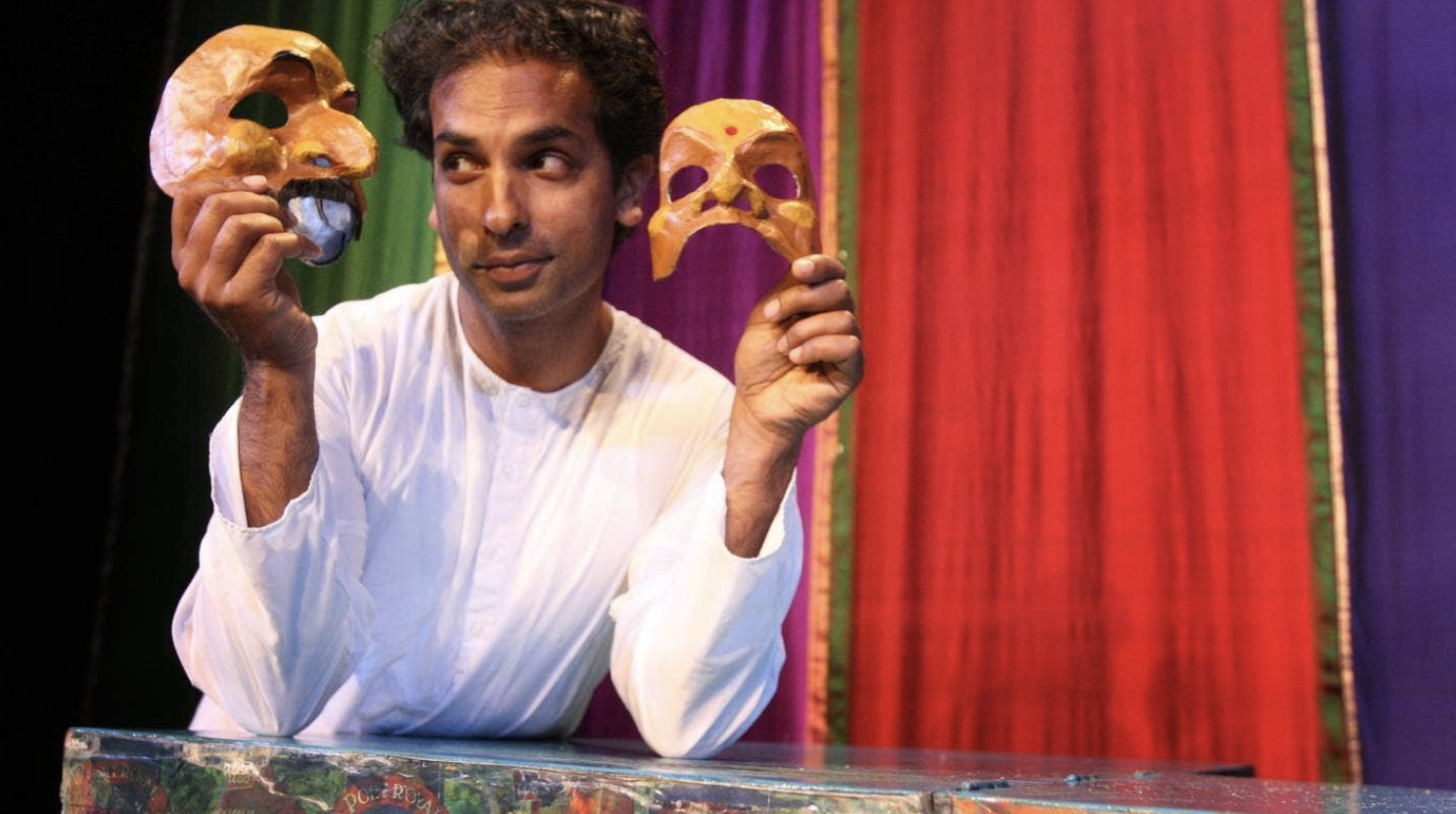 Man in white shirt holds two yellow theatrical masks, one smiling and one frowning, against colorful stage curtains.