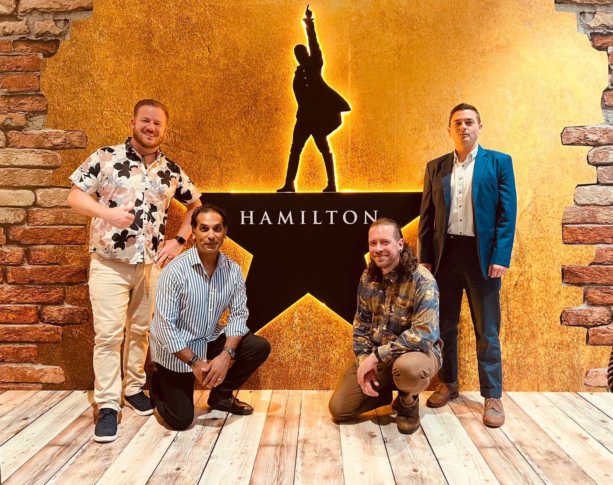 Four men posing in front of an illuminated Hamilton musical logo on a brick-and-wood backdrop.